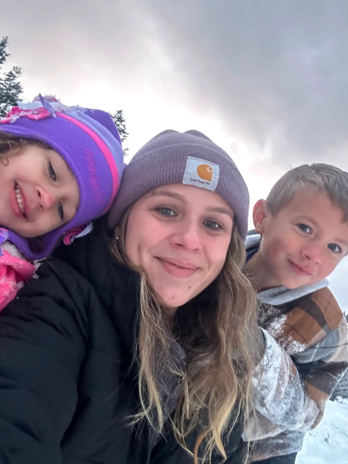 Joleene Souza in a beanie and winter jacket, smiling in the snow with her two kids, pine trees behind them